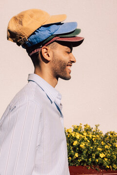 Side view of playful young man wearing multi colored caps against wall