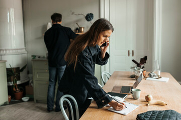 Female freelancer writing in diary while talking on smart phone at home