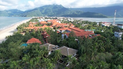 Fototapeta premium High-angle view of a coastal resort in Lang Co featuring red-tiled roofs, lush palm trees, sandy beach, and majestic mountains.