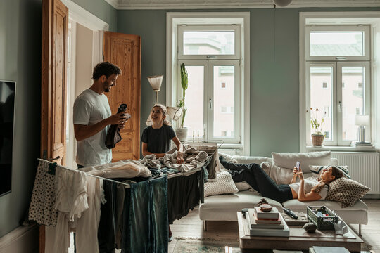Man folding clothes with daughter while woman using smart phone lying on sofa at home