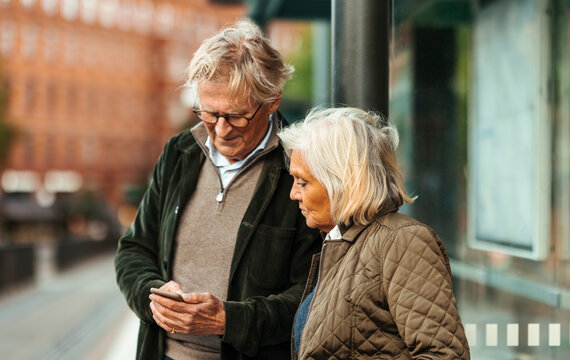 Senior woman looking at man using smart phone while standing at bus stop