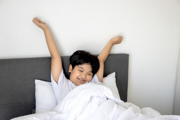 Asian boy waking up in bed, stretching his arms in the morning. Chubby young kid sleeping on...
