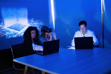 Group of young professionals working with technology in a modern office at night with blue lighting and virtual reality equipment