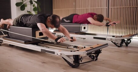 Two men perform pilates exercises on reformer machines, focusing on strength and technique in a well-designed fitness studio, enhancing their core stability and flexibility