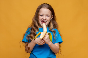 Happy funny laughing little girl without a baby tooth, wearing casual blue t-shirt eating ripe banana in her hands on yellow background, copy space. High quality photo. Children snack concept.