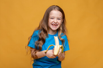 Happy funny laughing little girl without a baby tooth, wearing casual blue t-shirt holds ripe banana in her hands on yellow background, copy space. High quality photo. Children snack concept.