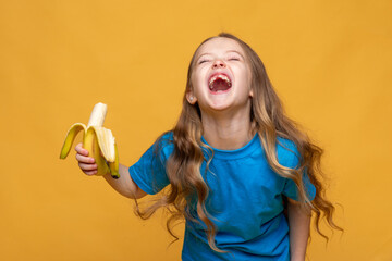 Happy funny laughing little girl without a baby tooth, wearing casual blue t-shirt holds ripe banana in her hands on yellow background, copy space. High quality photo. Children snack concept.
