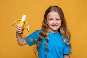 Happy funny laughing little girl without a baby tooth, wearing casual blue t-shirt holds ripe banana in her hands on yellow background, copy space. High quality photo. Children snack concept.