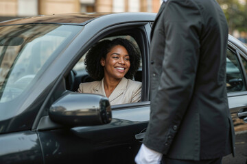 A black woman in a suit is smiling at a man who is standing outside her car. The man is wearing a suit and a white shirt