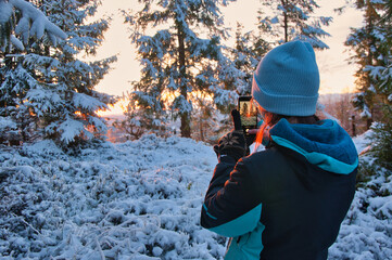 Kobieta fotografuje telefonem ośnieżony las o zachodzie słońca podczas zimowej przygody na świeżym powietrzu. © Pol Badger