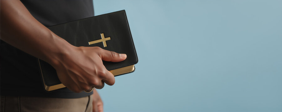 A Black man holding a Holy Bible with a gold cross on the cover. Christian faith, religion, and spirituality concept. Banner with copy space