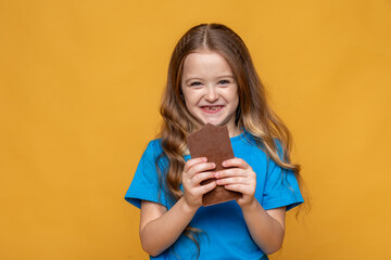 Cute little girl in blue t-shirt holding chocolate bar with nuts on yellow background, closeup. The concept of children's happiness and sweets addiction