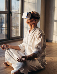 Calm elderly senior woman meditating in lotus position with virtual reality headset, experiencing peaceful technology for mindfulness and relaxation indoors