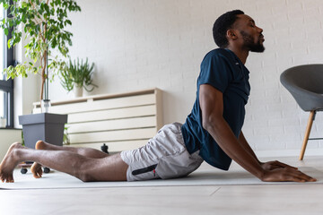 Naklejka premium African American man practicing yoga stretch at home in his living room, performing an upward-facing pose after workout. Indoor fitness scene focused on flexibility, recovery, calm and healthy lifesty