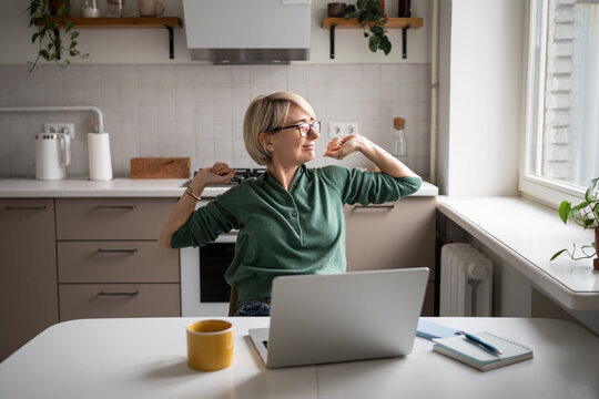 Smiling middle aged businesswoman stretching in chair during work on laptop, taking break to warm up, looks at window while working on remote from home in kitchen Female enjoys small break at work.