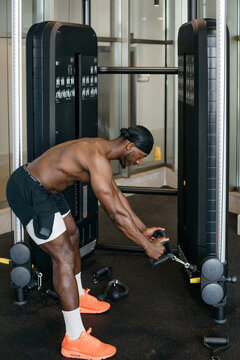 Athletic man training triceps with cable rope in gym. Muscular shirtless man leaning forward while performing cable rope pushdown exercise on strength machine indoors.