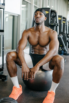 Athletic man resting on exercise ball in gym. Muscular African American athlete sitting on fitness ball during workout break at modern gym.