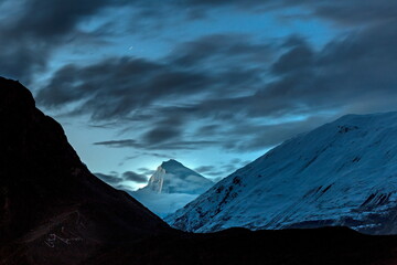 View of dark silhouettes of mountainous terrain frame a snow-dusted peak beneath a dramatic, clouded twilight sky, Hunza, Gilgit Baltistan, Pakistan.