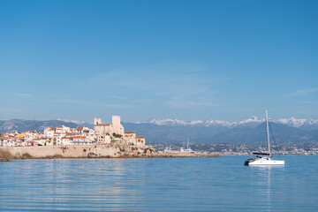 Sailboat on Calm Mediterranean Sea with Coastal City and Snowy Mountains in Background