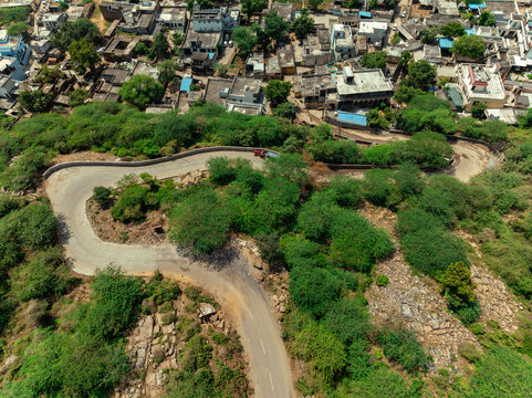 Aerial view of a winding road snaking through dense green foliage towards the clustered buildings of a village, Mangar, Haryana, India.
