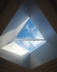 Triangle skylight in indoor ceiling: glass panel reveals blue sky with white clouds, white architectural structure, natural sunlight streaming in, modern interior design element with bright open view.
