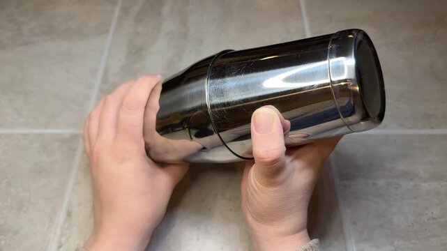 Female hands assemble a metal cocktail shaker by placing both halves together, preparing for mixing a drink. Minimal overhead view with a clean background and hands-only action.