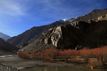 View of rugged mountains rise dramatically above a serene river lined with vibrant red trees under a crisp blue sky, Passu, Gilgit Baltistan, Pakistan.