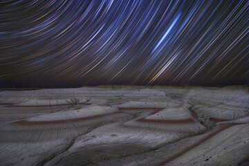 View of celestial trails dance above the arid landscape, painting the night sky with streaks of light over the red and white textured ground, Senek, Mangystau Region, Kazakhstan.