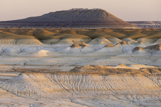 View of the layered, undulating terrain under a soft, dawn sky, with a mesa rising in the distance, casting shadows across the textured landscape, Senek, Mangystau Region, Kazakhstan.