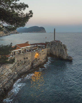 View of a restaurant perched on rugged cliffs where the Adriatic Sea kisses the shore as dusk paints the sky in watercolor hues, Petrovac, Budva Municipality, Montenegro.