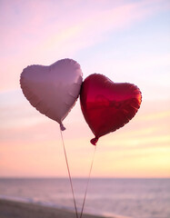 Fototapeta premium Two heart-shaped balloons float gently against a soft pastel sunset sky over the ocean