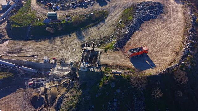 Beneath a bright sky, construction vehicles move through a busy site filled with gravel and machinery. Shadows stretch across the ground as work continues in the late afternoon sun.