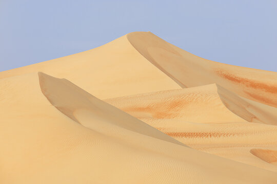 View of golden dunes sculpted by wind under a pale blue sky, creating a serene desert landscape of textured sand and subtle color gradients, Bateen Liwa, Abu Dhabi, United Arab Emirates.