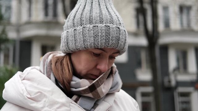 Portrait of mature woman in wool hat standing on the urban street with mobile phone in hand. Middle-aged 40s female using smartphone, walking the city at cold autumn day. Living in modern district