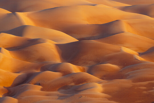 View of undulating sand dunes, painted in warm, fiery hues under the desert sun, creating a mesmerizing pattern of light and shadow, Bateen Liwa, Abu Dhabi, United Arab Emirates.