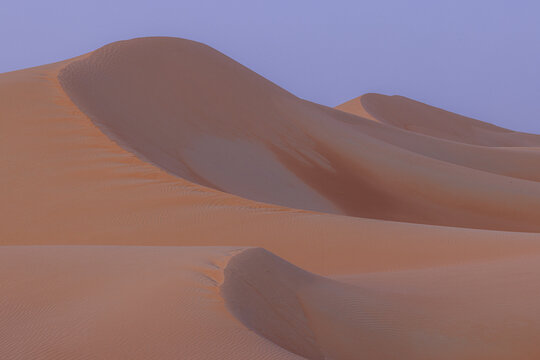 View of rolling dunes of orange sand ripple under a soft lavender sky at dusk, creating a serene desert landscape, Bateen Liwa, Abu Dhabi, United Arab Emirates.