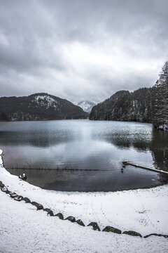 View of the serene lake mirroring the clouded sky, bordered by snow-covered banks and dark, dense forests, creating a tranquil winter scene, Bavaria, Germany.