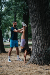 Fototapeta premium A man helps a woman perform a handstand against a large tree in a park. They practice balance and support in an outdoor fitness moment.