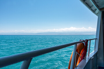 Fototapeta premium A view from a boat or ferry of a large body of water with mountains in the distance and a clear blue sky. A metal railing and an orange lifebuoy are visible in the foreground. 