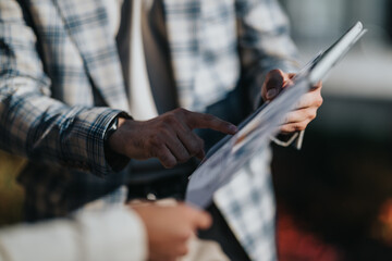 A business professional uses a tablet to review documents. Two people discuss and point at a brochure in a casual outdoor setting.