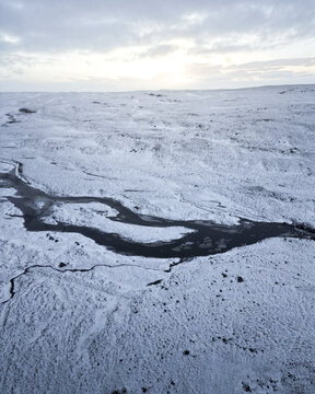 Aerial view of a serpentine river carves its path through the stark, snow-covered plains under a pale sky, creating a striking contrast, Vik, Western Region, Iceland.