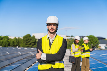 Male engineer standing with crossed arms over solar panel field. Team members working in background, focused on sustainable technology