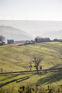 View of rolling green hills dappled with shadows leading to a building, with wind turbines on the horizon, all under a hazy sky, Trier, Rhineland Palatinate, Germany.
