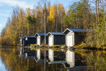 Row of modern dark grey boathouses by a calm lake in autumn, golden forest trees reflecting in water, scandinavian style lakeside architecture