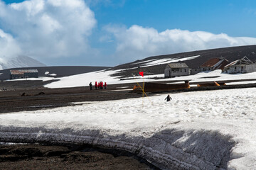 View of stark black volcanic sands meeting icy white snowdrifts under a cloudy sky, with distant buildings and a cluster of people in red, Deception Island, Antarctica.