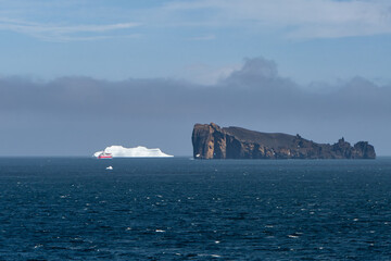 View of a red cruise ship sails past a stark white iceberg and a dark, rocky island under a hazy sky, contrasting with the deep blue sea, Deception Island, Antarctica.
