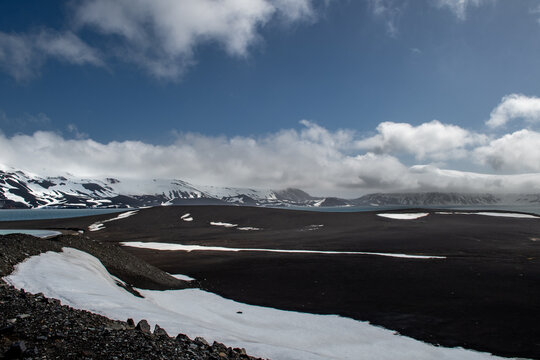 View of stark black volcanic sands meet icy patches under a vast sky, contrasting the rugged terrain with the serene horizon, Deception Island, Antarctica.