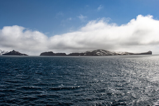 View of the dark, choppy sea contrasting with the snow-dusted islands under a bright, partly cloudy sky, Deception Island, Antarctica.