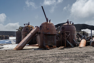 View of decaying, rusty industrial tanks and debris scattered across the desolate landscape under a cloudy sky, Deception Island, Antarctica.