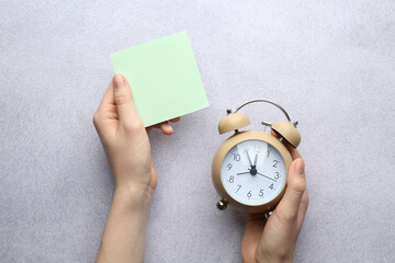 Woman with sticky note and alarm clock at light grey table, top view